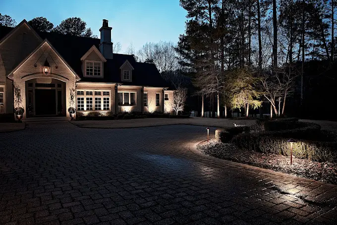 A large house with dormer windows is beautifully lit by residential lighting in Jefferson County, AL, during the early evening. Surrounded by trees, its cobblestone driveway features landscaped bushes illuminated by elegant pathway lights beneath the deep blue sky.
