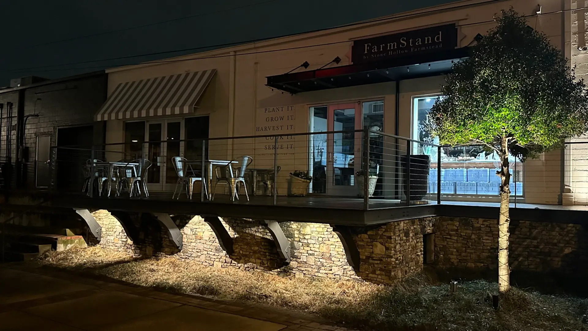 Night view of a restaurant with a patio featuring metal chairs and tables beneath enchanting outdoor lighting. A tree adorned with lights stands on the elevated stone pillars. The restaurant has a striped awning over a window and a lit sign above the entrance, capturing Jefferson County, AL charm.