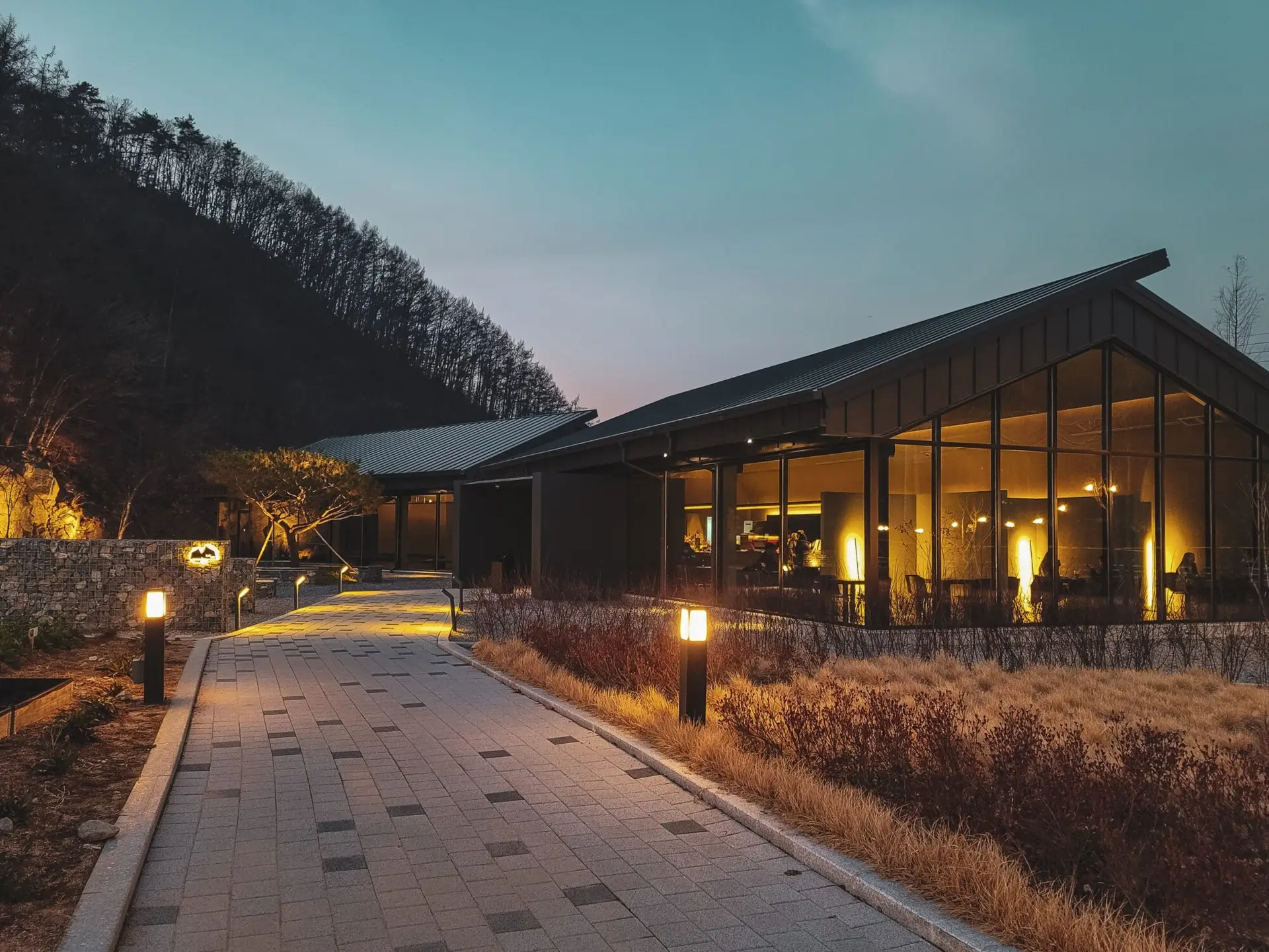 A modern building with large glass windows is illuminated at dusk, showcasing exceptional outdoor lighting in Jefferson County, AL. A stone path leads to the entrance, surrounded by dry grass and shrubs. Trees and a hill are visible in the background under a twilight sky.