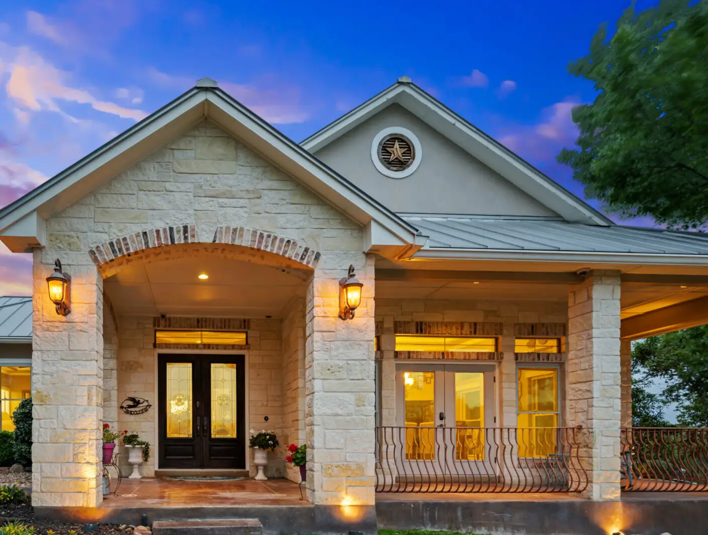 A charming stone house with a wide porch is illuminated at dusk, featuring elegant outdoor lighting. The front boasts arched windows and a double door flanked by lantern-style lights. Set against a soft blue sky with clouds, greenery frames the entrance in Jefferson County, AL.