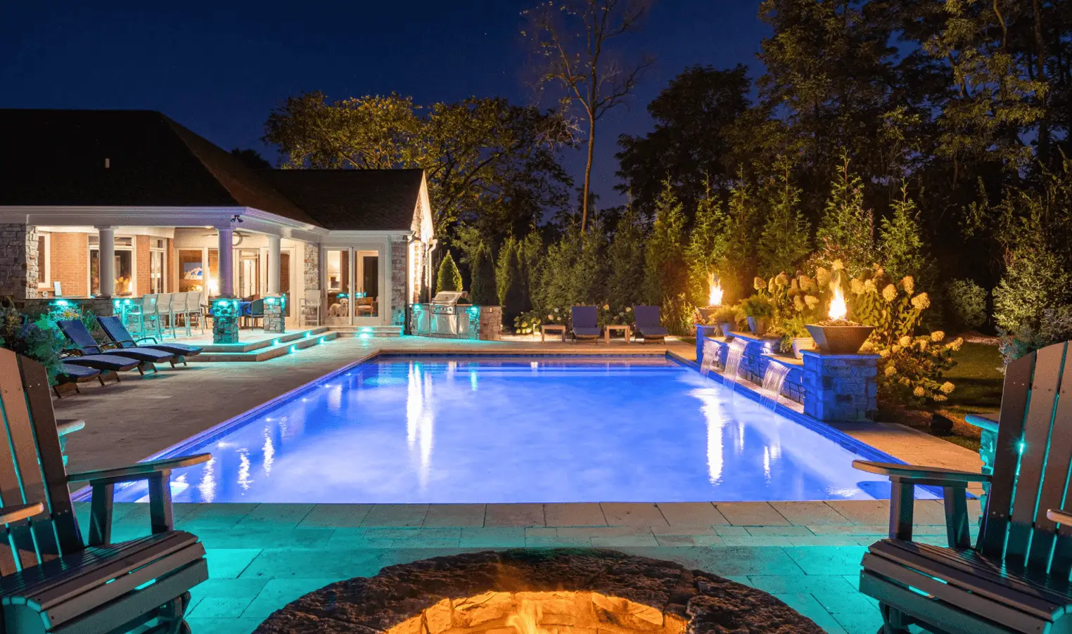 An illuminated backyard pool at night with a modern house in the background, enhanced by exquisite outdoor lighting. The pool, surrounded by lounge chairs and decorative fire pits, creates a cozy atmosphere. Lush trees and shrubs provide natural borders to this Jefferson County AL oasis.
