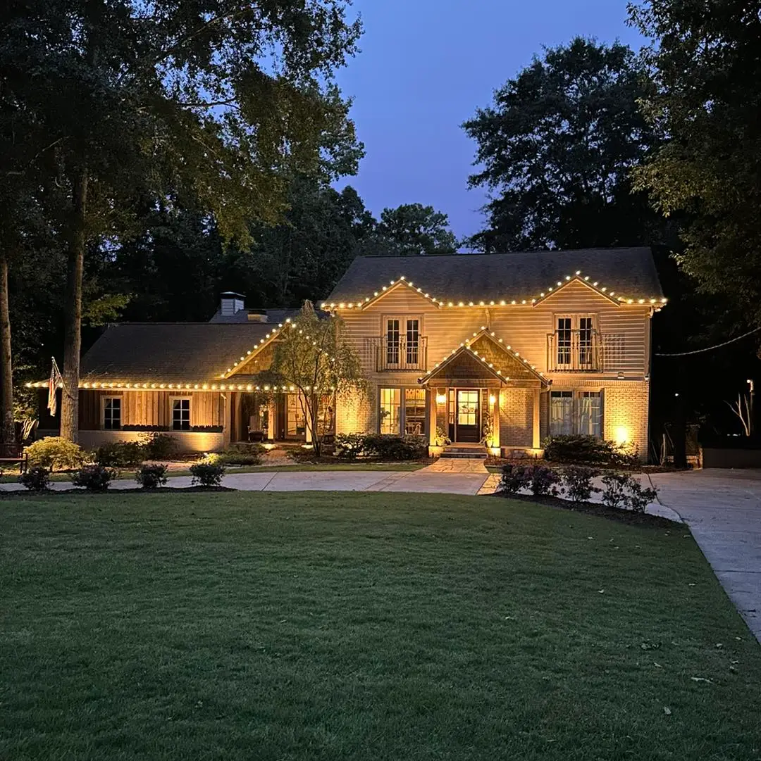 A two-story house in Jefferson County AL, highlighted by warm white string lights, has its roof and porch beautifully outlined at twilight. The inviting residential lighting showcases lush greenery, including a vibrant lawn and tall trees, while the driveway gracefully leads to the front door.