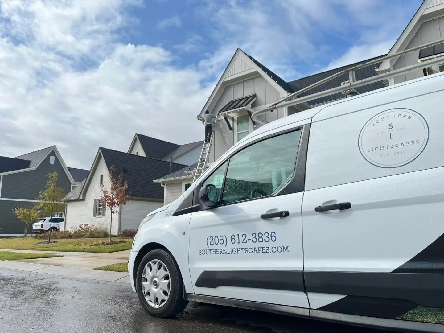 A white van labeled "Southern Lightscapes" is parked on a residential street in Jefferson County, AL. A person on a ladder is busy installing festive residential lighting on a house. The sky is partly cloudy, and the leaves are beautifully changing colors.