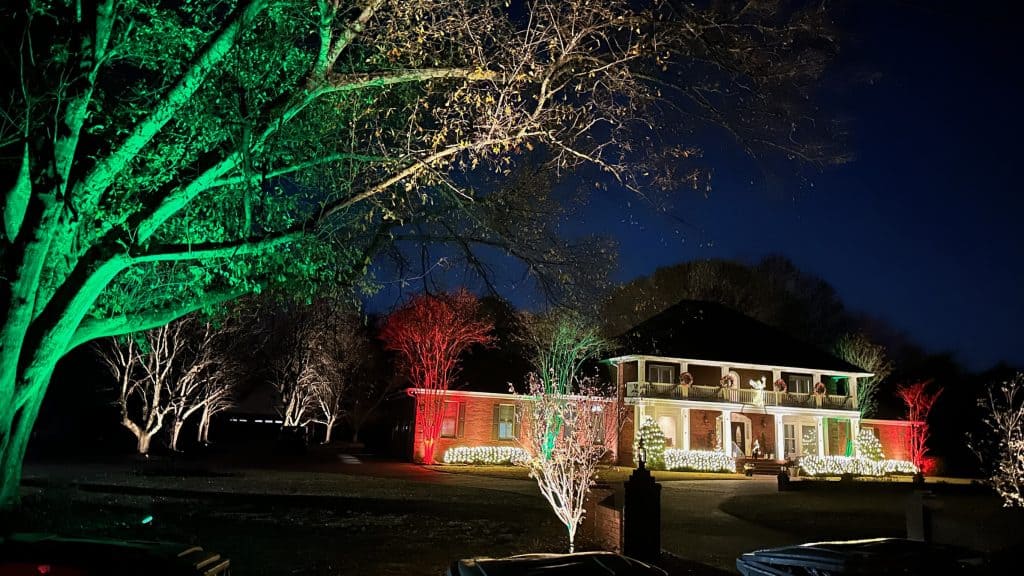 A two-story house in Jefferson County, AL, is adorned with vibrant outdoor lighting for the holidays. The façade glows with red and green lights, and the trees in the yard shimmer in similar hues. The scene is beautifully set against the night sky.