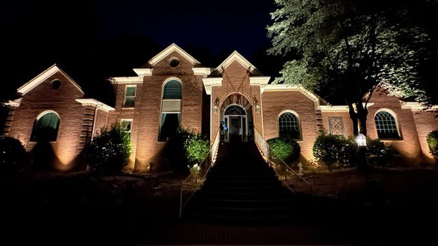 A large brick house in Jefferson County, AL, is beautifully illuminated by outdoor lighting at night. It features tall windows, a central archway, and multiple gabled roofs. The building is framed by trees and shrubs, with stairs leading up to the entrance.