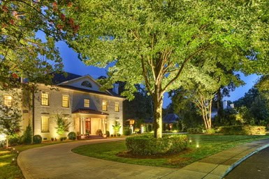 Nestled in Jefferson County, AL, this large two-story house is surrounded by trees and a landscaped lawn. Warm outdoor lighting illuminates its exterior as a curved driveway leads to the column-framed entrance, all set against a twilight sky.