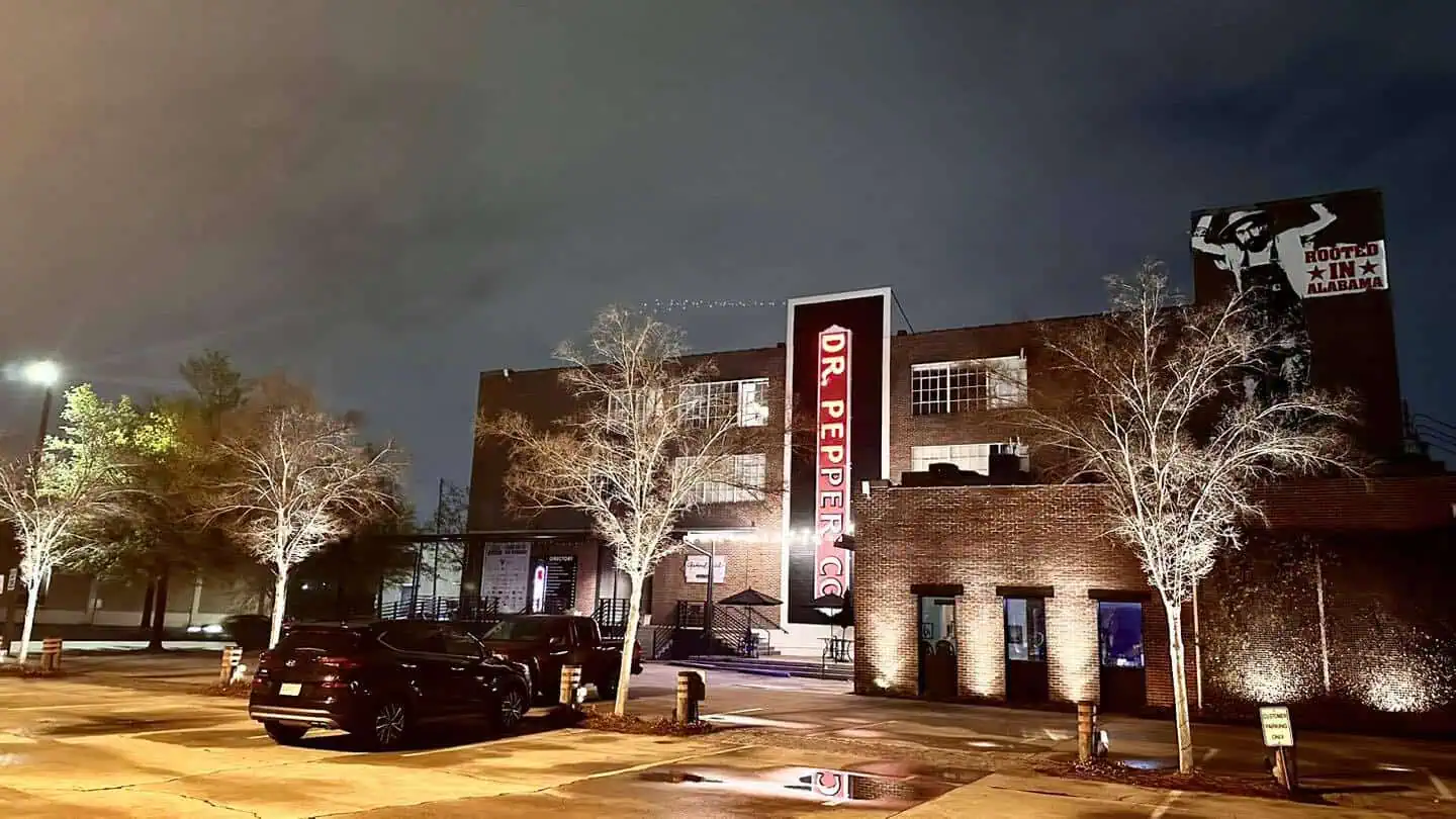 Night view of the Dr. Pepper building with prominent red signage, surrounded by bare trees and parked cars. A mural with a star is visible on the right side of the building. The sky is overcast, casting a soft glow.