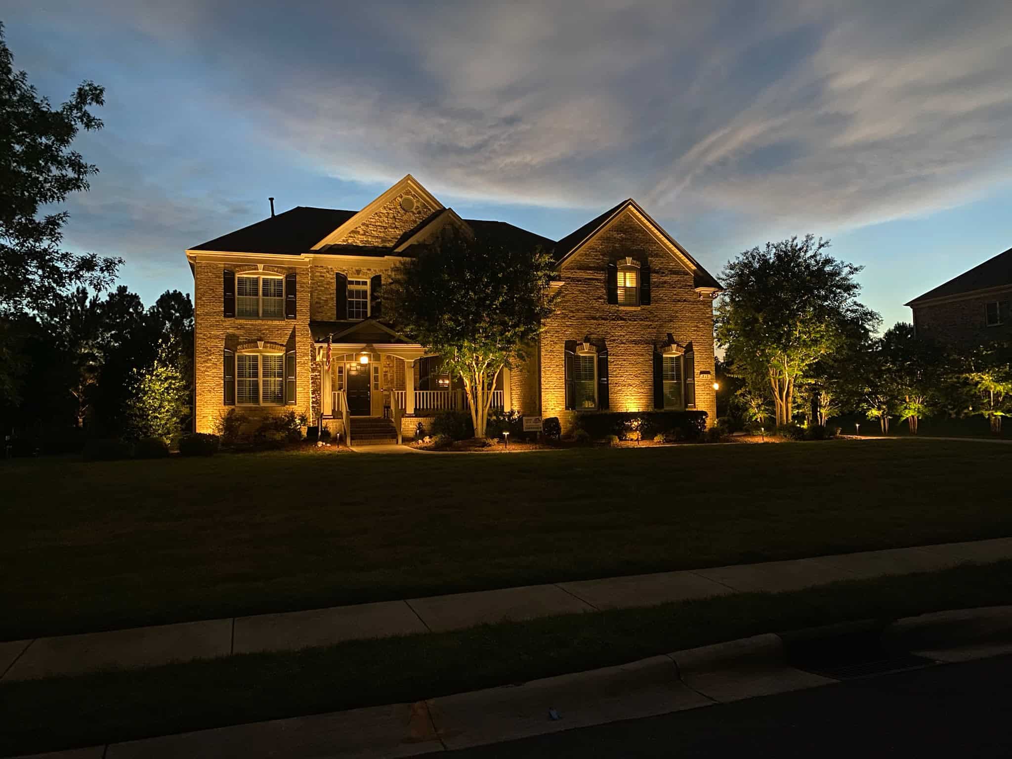 A large brick house with columns is beautifully illuminated by outdoor lighting at dusk. Nestled in Jefferson County, AL, the front yard dazzles with neatly trimmed bushes and trees, set against a dramatic sky of dark clouds and fading light.