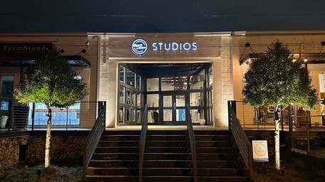 A building entrance at night, with "Studios" glowing above, showcases vibrant outdoor lighting. Flanked by two illuminated trees, the stairs lead to large glass doors and stylish wooden siding—a modern architectural gem in Jefferson County, AL.