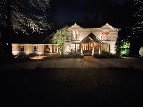 Illuminated two-story house at night in Jefferson County, AL, with warm outdoor lighting accentuating its wooden facade. The house features two small balconies, surrounded by trees, with a driveway leading up to the entrance.
