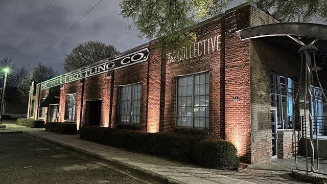 A brick building with large windows is illuminated by outdoor lighting at night. The facade reads "Ice & Bottling Co." and "Collective" as a streetlamp casts a greenish glow on the sidewalk lined with bushes in Jefferson County, AL.