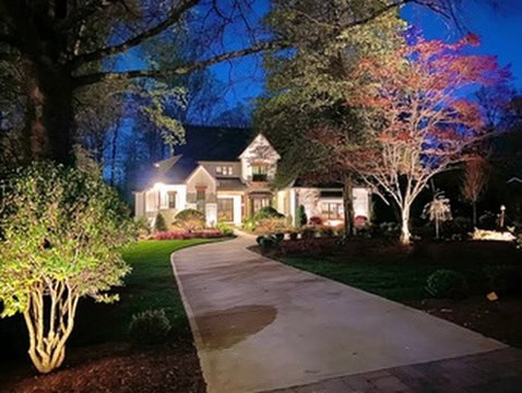 A warmly lit house with a gabled roof, nestled in Jefferson County, AL, is framed by trees and landscaped gardens at dusk. The long driveway, adorned with outdoor lighting and flanked by illuminated shrubs and a crimson-leaved tree, guides you to the inviting entrance.