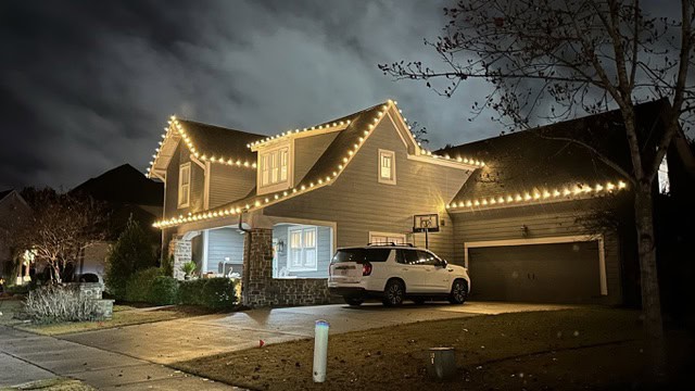 A two-story house in Jefferson County, AL, is adorned with glowing white string lights along the roof. A white SUV is parked in the driveway near a basketball hoop. The sky is cloudy, and bare trees are visible, suggesting a winter scene enhanced by charming outdoor lighting.
