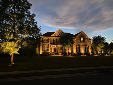 A well-lit two-story brick house at dusk in Jefferson County, AL, features dramatic outdoor lighting that highlights trees and architectural features. The sky is subtly illuminated by the remaining light from sunset, enhancing the serene ambiance.