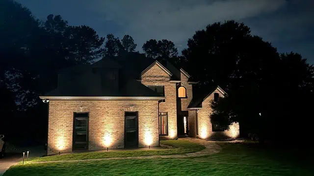 A two-story brick house in Jefferson County, AL, is beautifully lit at night with outdoor lighting placed along the ground, accentuating its facade. A curved pathway leads to the entrance, while tall trees form a dark silhouette in the background.