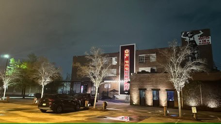 Exterior view of a building at night in Jefferson County, AL, showcasing a prominent vertical "Dr Pepper" sign. The brick structure glows with outdoor lighting and a mural on the side. Several cars are parked in the foreground, illuminated by streetlights.