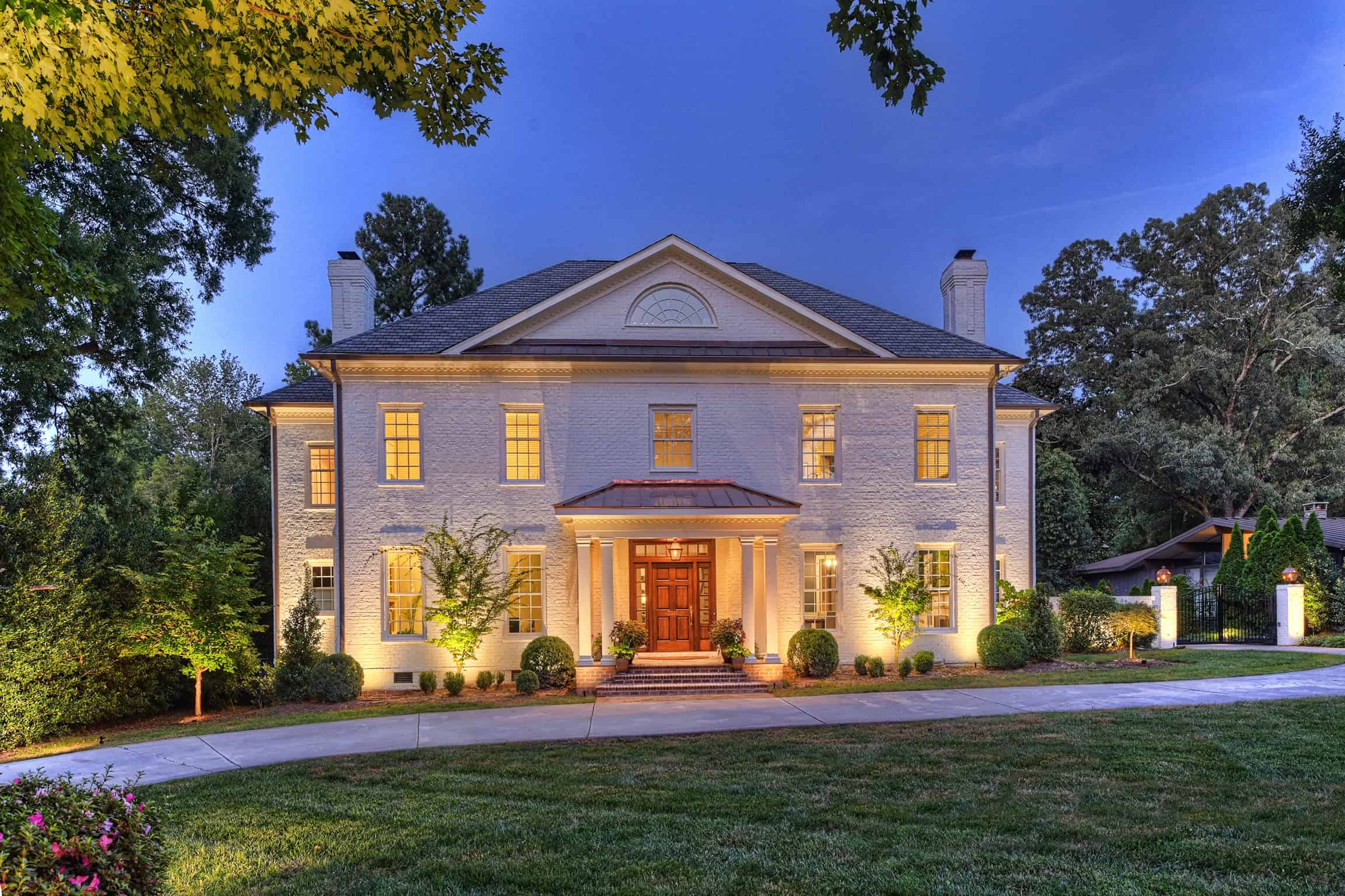 A grand, two-story colonial-style house at dusk in Jefferson County, AL, features a white brick exterior, symmetrical windows, and a central porch with columns. Warm outdoor lighting elegantly illuminates the facade while a curved driveway is embraced by well-kept greenery.