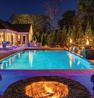 A luxurious backyard at night showcases a lit swimming pool, a circular fire pit, and a well-lit house with large windows. Enhanced by Jefferson County, AL's finest outdoor lighting, torches and trees envelop the area, crafting an inviting and cozy atmosphere.
