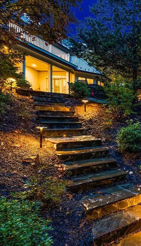 Stone steps, lit by outdoor lighting, guide the way up a slope to a warmly illuminated porch. Nestled in nature's embrace with trees and shrubs, this charming setting in Jefferson County, AL comes alive under the night sky.
