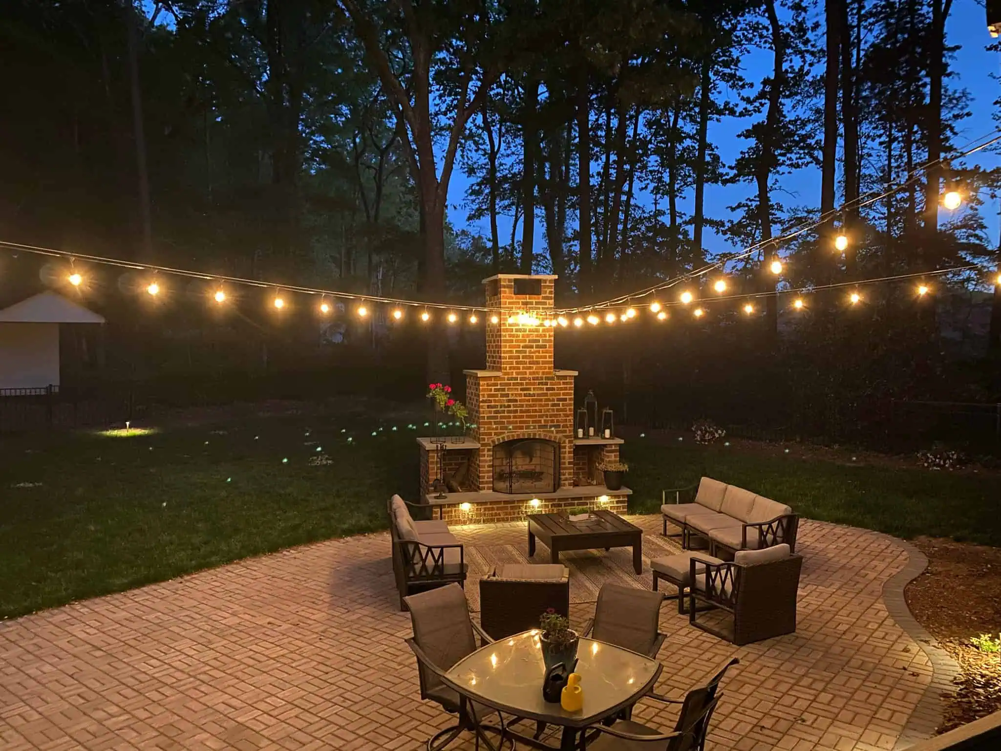 A cozy outdoor patio in Jefferson County, AL, glows at dusk with charming outdoor lighting. String lights hang above a brick fireplace surrounded by cushioned patio furniture, along with a dining table and chairs set against a backdrop of lush greenery.