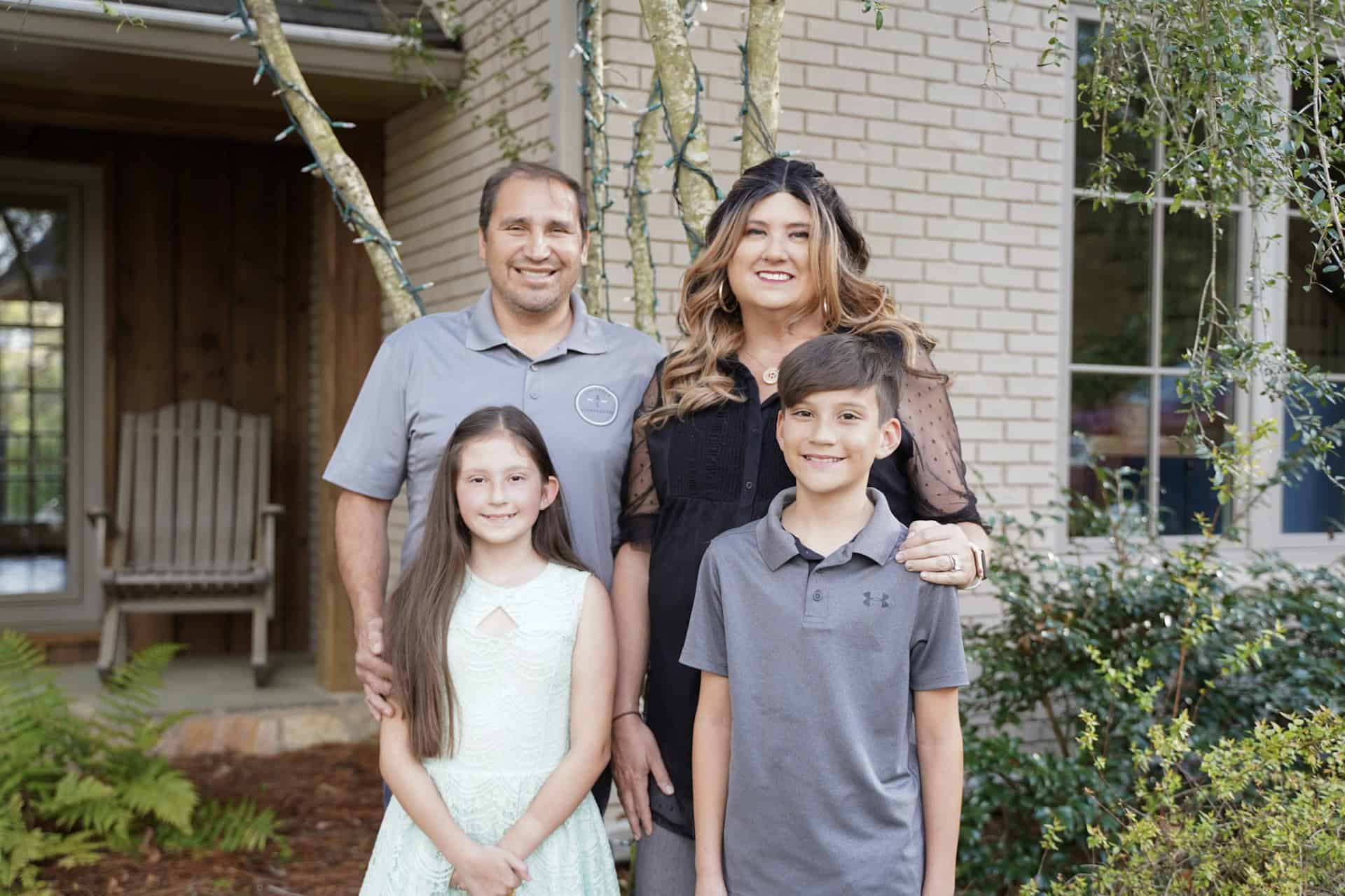 A family of four stands together outside a house. A man and woman are behind two children. The man and boy are wearing gray shirts, while the woman is in black and the girl is in a light dress. Trees and shrubs are in the background.