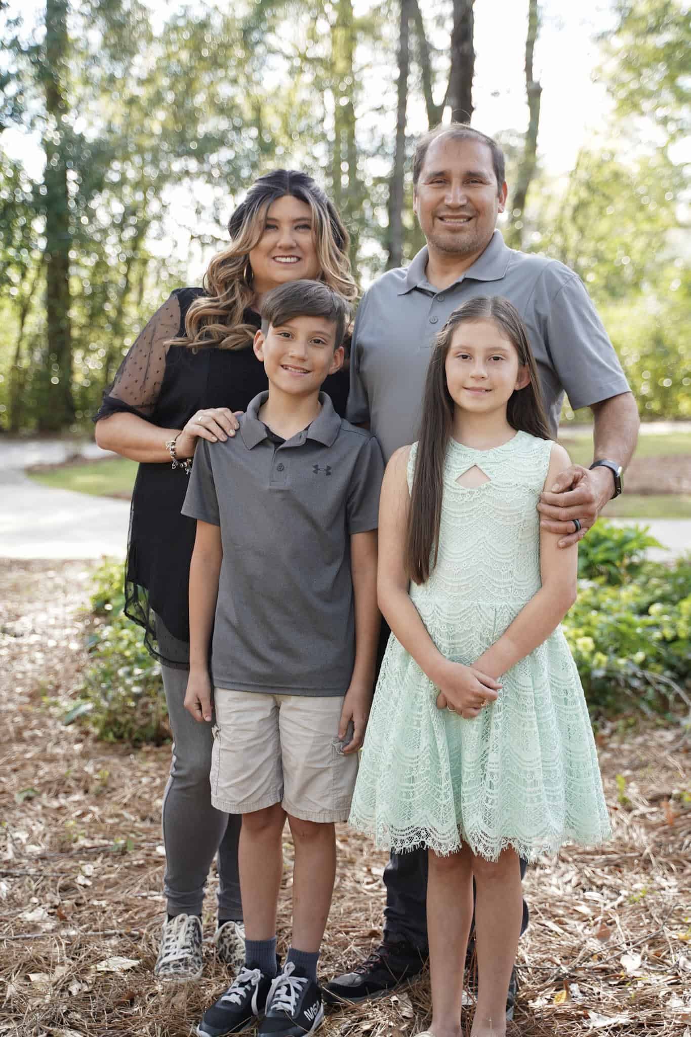 A family of four poses outdoors on a sunny day. The woman stands beside the man, both smiling. In front of them, a boy in a polo and shorts and a girl in a light green dress smile. Trees and a path are in the background.