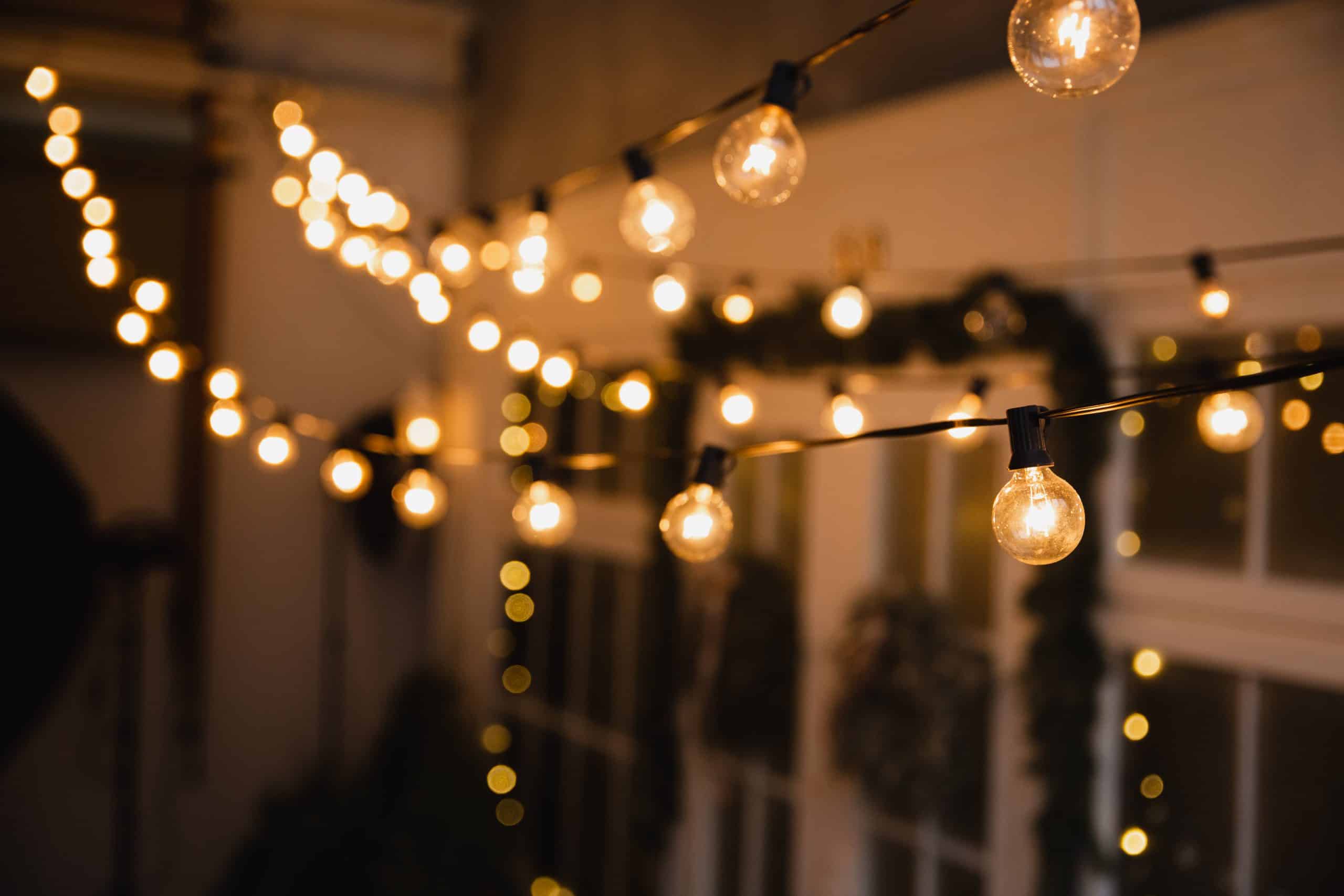 String lights with warm glowing bulbs are hanging in an indoor setting, reminiscent of festive outdoor lighting found in Jefferson County, AL. The background is softly blurred, showing decorative greenery and soft lighting, creating a cozy, inviting atmosphere.