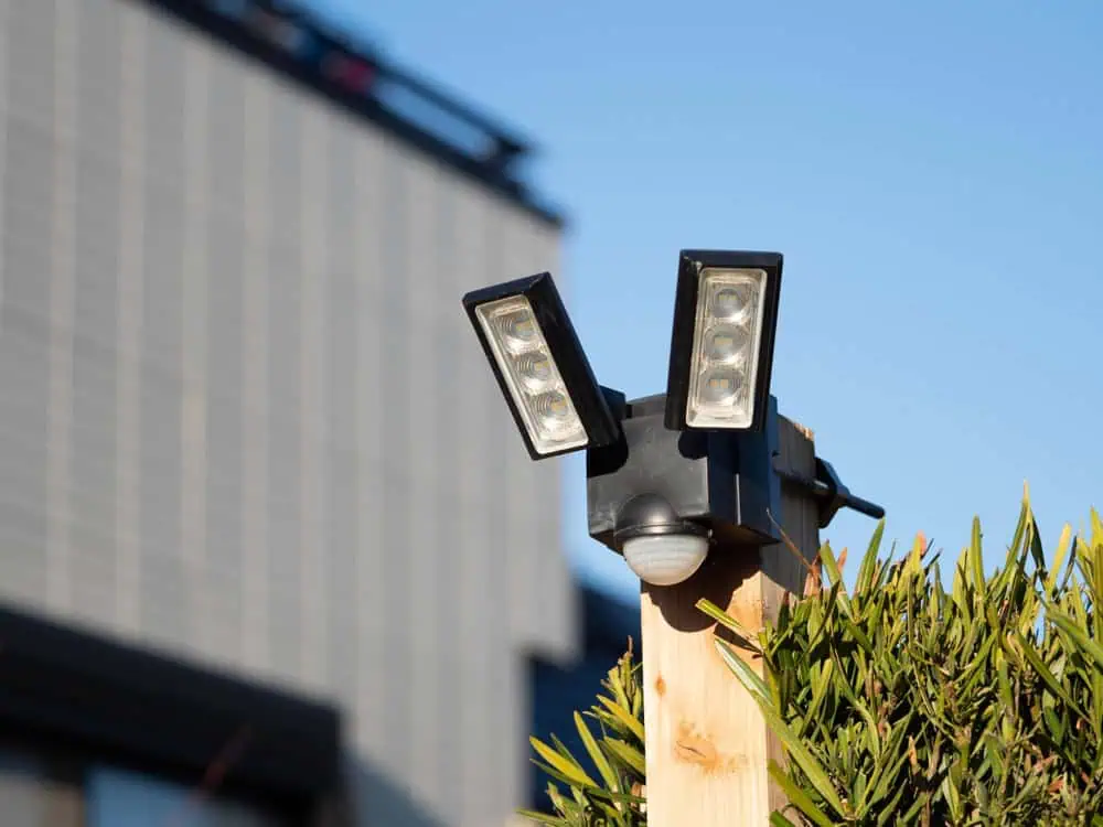 A security camera with dual rectangular lights mounted on a wooden post beside some shrubs. A modern building with vertical lines is visible in the background against a clear blue sky.
