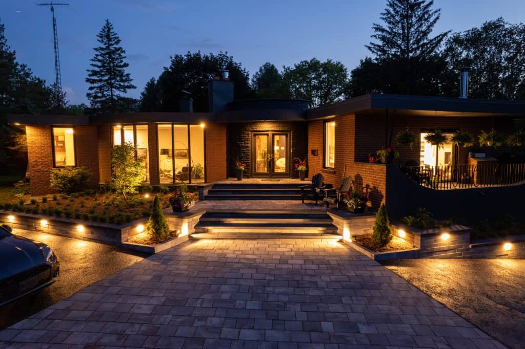 In Jefferson County, AL, an illuminated modern brick house at dusk features large windows and a landscaped yard. The outdoor lighting warmly accentuates the steps and entrance, while trees are silhouetted against the evening sky. A car rests on the paved driveway.