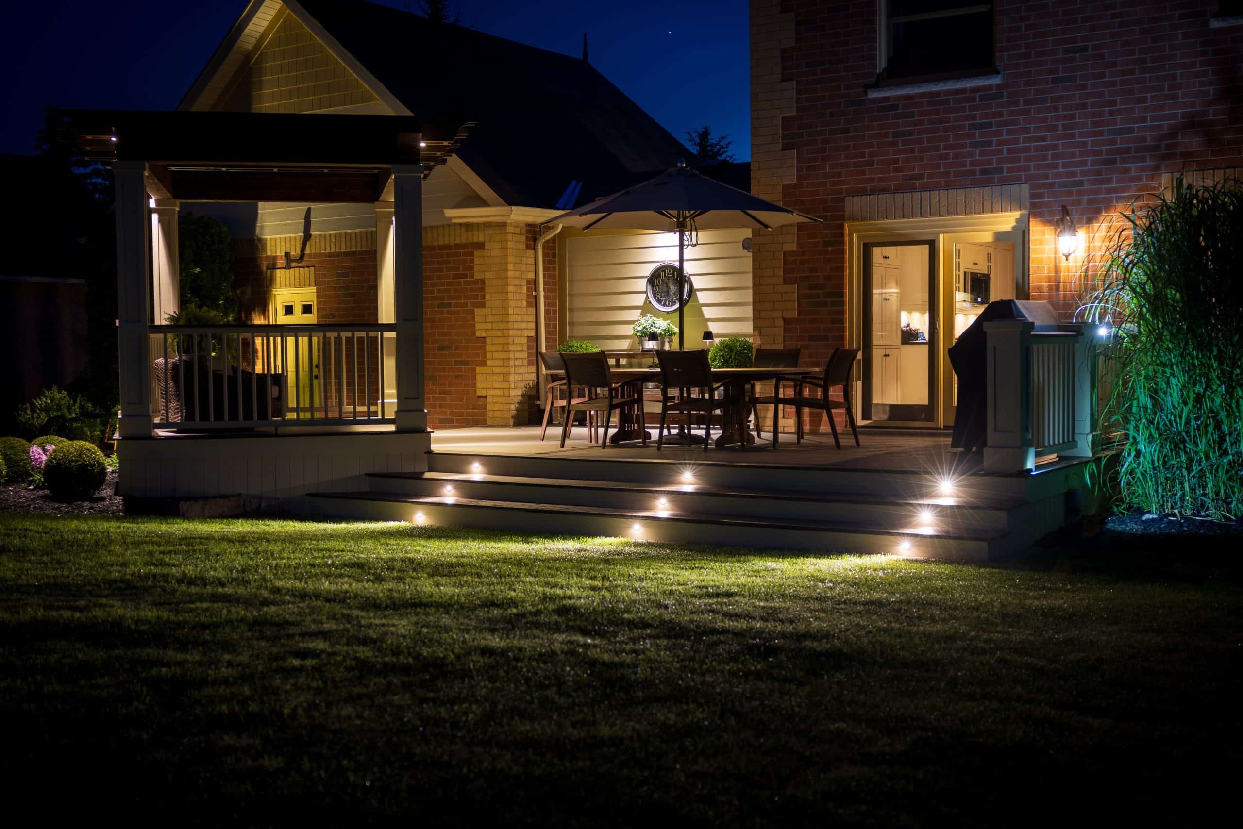 A backyard patio at night with warm outdoor lighting creates an inviting ambiance. In Jefferson County, AL, a table and chairs rest under a large umbrella on the wooden deck. Soft lights illuminate the stairs leading to the lawn, while a brick house and gazebo frame this cozy oasis.