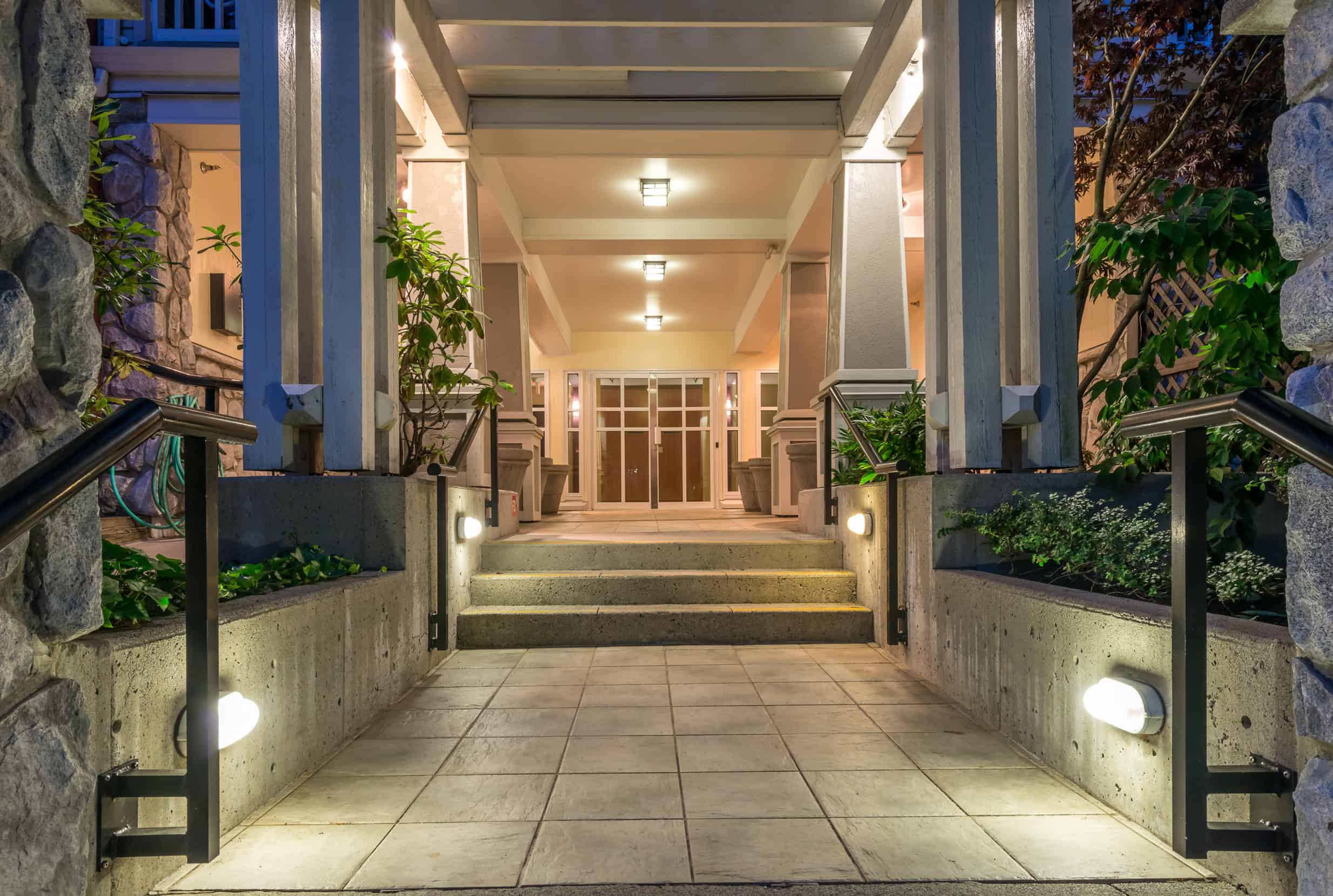 The building's entrance boasts well-designed outdoor lighting, creating a welcoming glow along the walkway. Lined with lush greenery in Jefferson County, AL, the path leads to a doorway under a covered porch supported by pillars, with warm lights enhancing its charm.