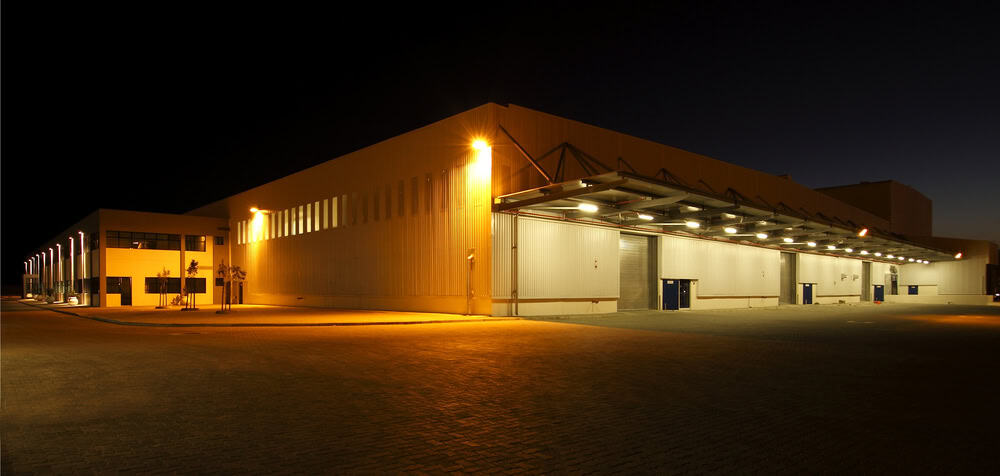 A large industrial warehouse is illuminated at night. The building features multiple loading bays on the right, with lights casting a warm glow on the exterior. The sky is dark, and the area appears quiet and deserted.