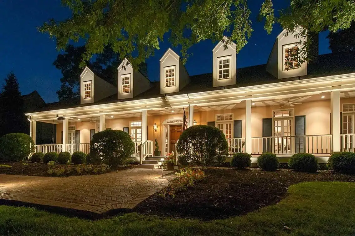 A large, well-lit house at night with three dormer windows, a wide front porch, columns, and neatly trimmed bushes—perfectly illuminated by expert lighting services in Jefferson County, AL.