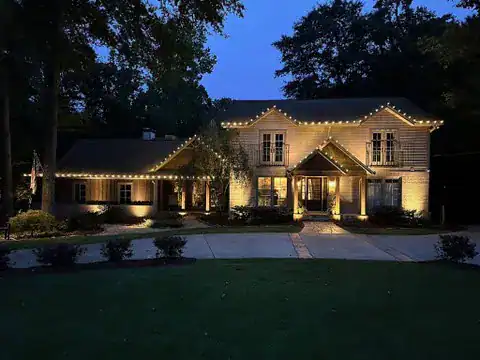 A two-story house in AL with warm exterior lights and string lights outlining the roof, surrounded by trees and a neatly trimmed lawn, photographed at dusk—showcasing expert lighting services Jefferson County residents trust.